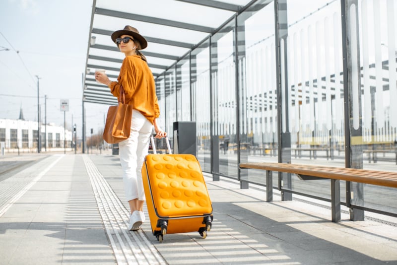 年轻的女性旅行者带着一个黄色的行李箱走在现代交通站外，后视图。